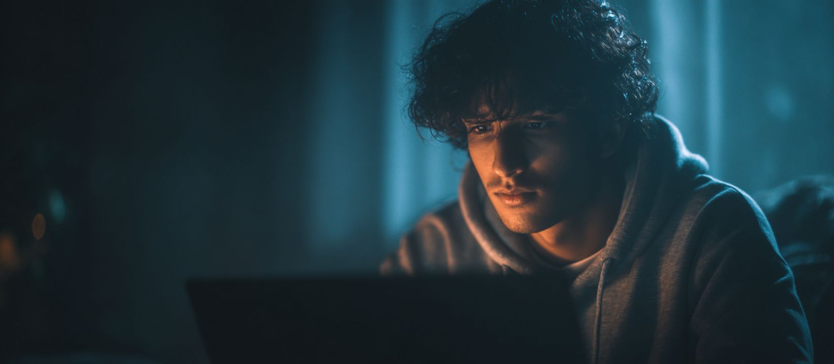 A man in a hoodie sits in front of a laptop playing Aviator in a dark room with soft blue light